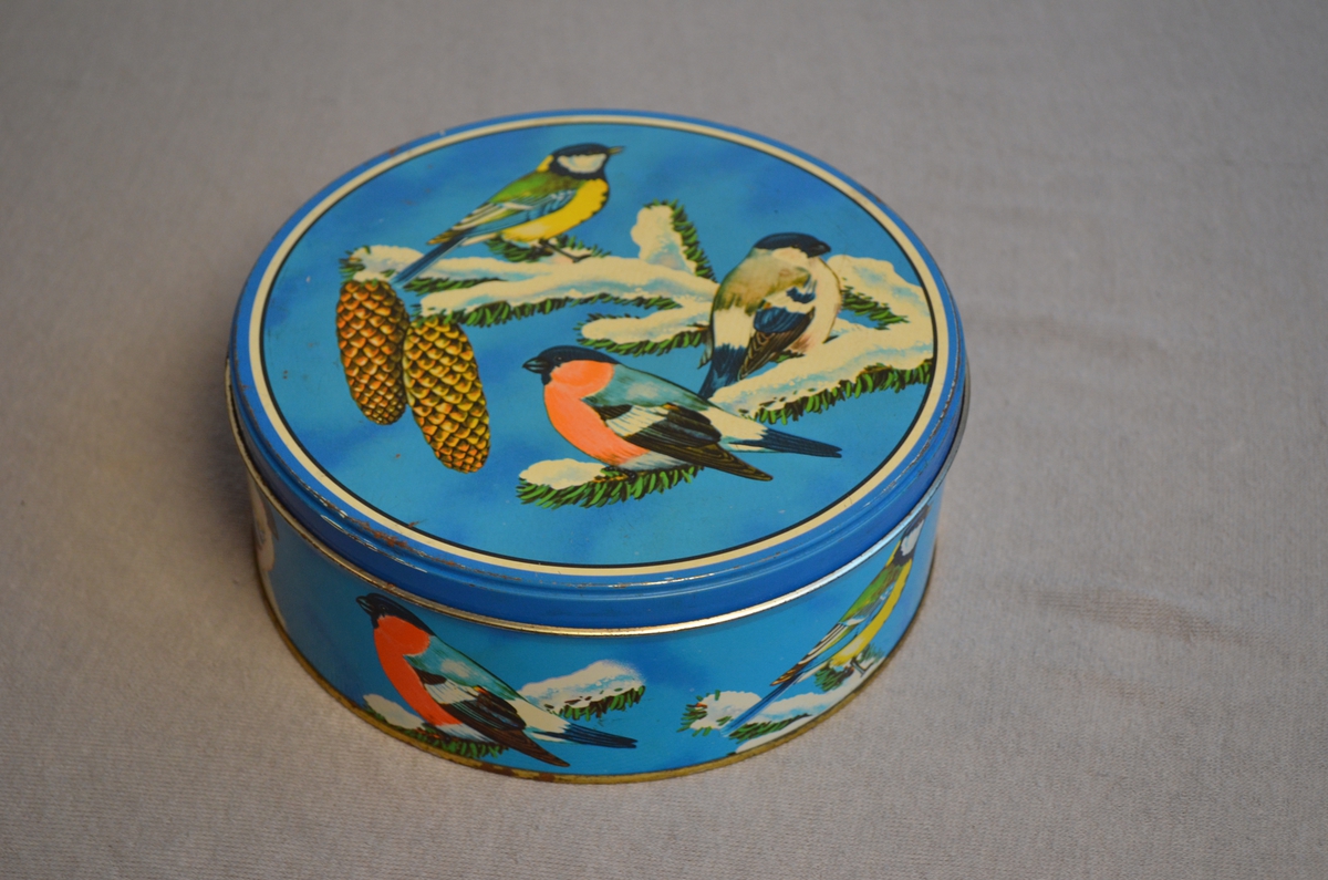 Rund kakeboks. Blå med motiv av småfuglar som sit på ei grein (grantre). Det ligg framleis kromkaker i boksen.