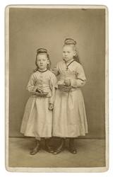 To barn, jenter, i finkjole med kurv og oppsatt hår, knappe 