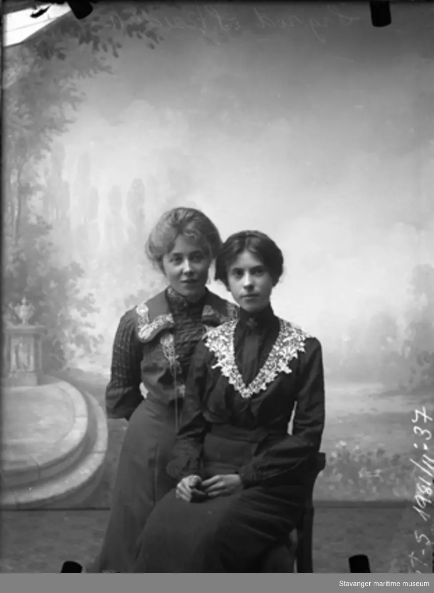 Portrett. Bakteppe med svingtrapp og hagemotiv. To unge kvinner, en sitter, den andre står. Mørke kjoler med høy hals, nupperellekrager. Fra fotograf Bergs atelie i Florø, 1904-1908.
