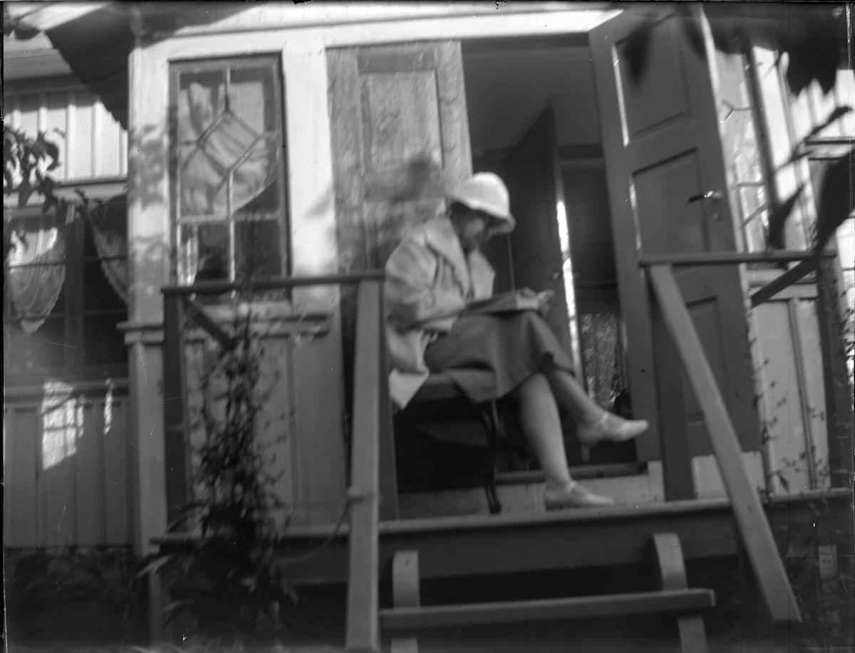 'Bildtext: ''Kyrkesund 1929.'' :: 1 kvinna sittande på en stol på veranda till hus, läser en bok. ::  :: Ingår i serie med fotonr. 5233:1-14'