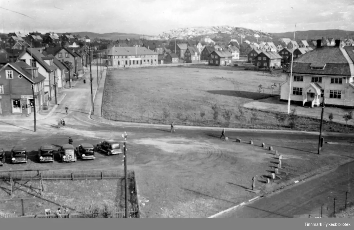 Parti fra Kirkenes. Viulsgate med A/S Sydvarenger sin administrasjonsbygning til høyre. Drosjeholdeplass i forgrunnen der det står flere biler parkert i rad. En av bilene er en 1935-36-modell så bildet må være tatt på denne tiden eller senere.