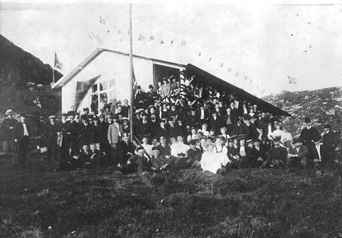 Større gruppe foran fjellhytte, taket er smykket med flagg. Innvielse av fjell-logen Harald`s hytte Kringsjå i 1909.