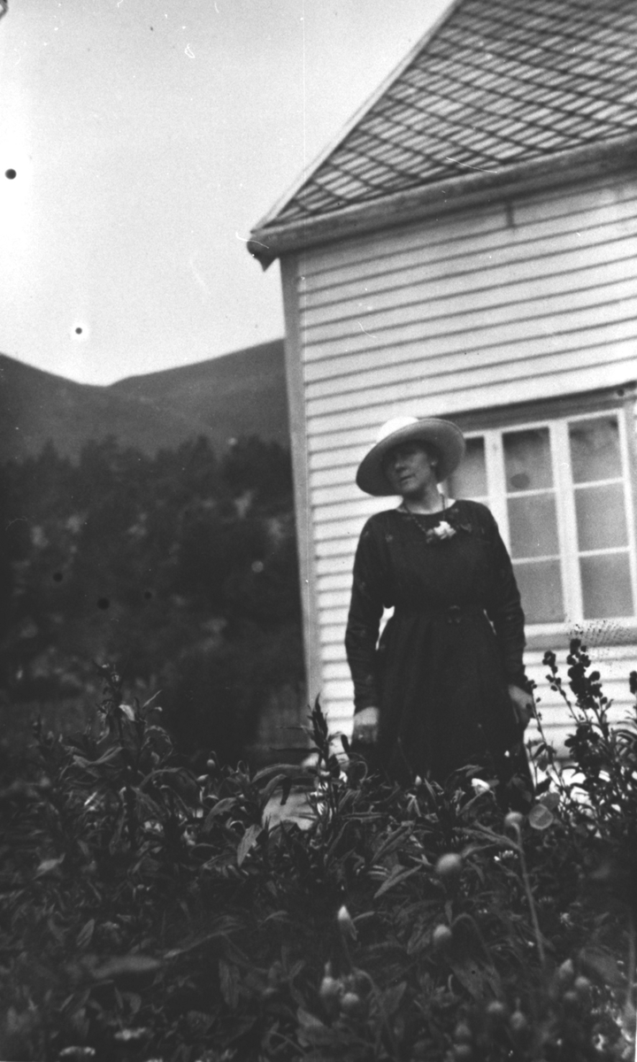 Dame med hatt i hagen foran et hus.