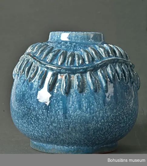 Klotrund vas med dekor av pålagd bladsinga i relief. Vit engobe med blå glasyr. I botten handskriven signatur: Greta