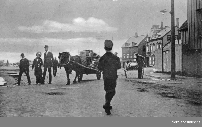 Gatebilde ved Jakhellngården. Barn og voksne. 1 hest med kjerre og en mannsperson med ei håndkjerre. Bebyggelse t.h., Grand hotell.
Se NM.F.001132-00001