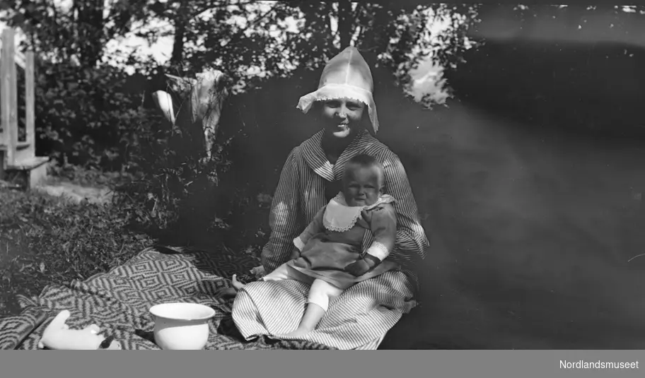 En kvinne som sitter på et pledd med en unge, Unni Strand, f. 1917 på fanget. Kvinnen har en slags hette på hodet. På pleddet ligger en lekehund, og til høyre for den ståre en potte.