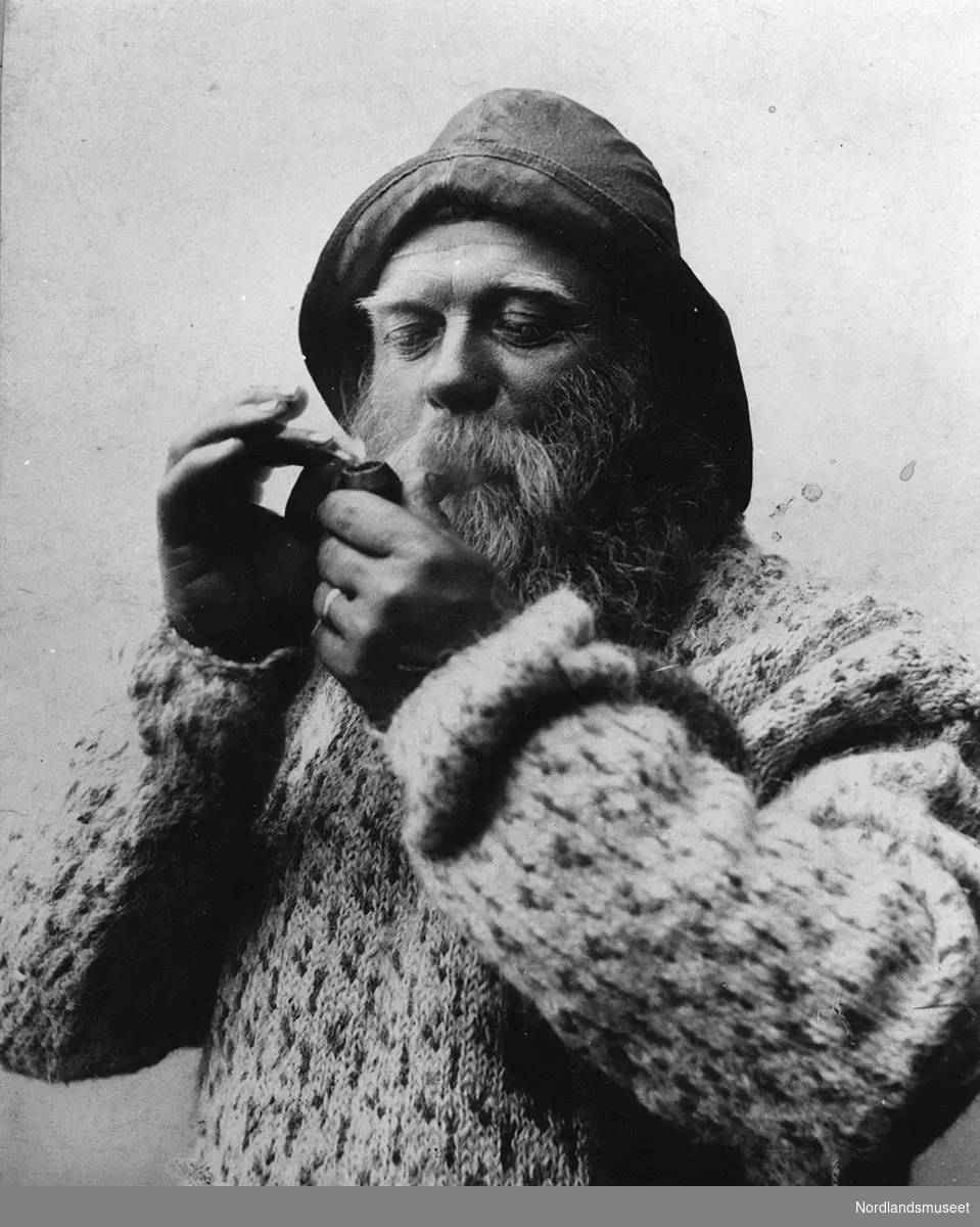 Portrett av mann med sydvest, skjegg og genser. Johannes Folgerø tenner på pipa. Bildet var et av de som ble tatt av ham i anledning fiskerimuseets åpning 1904.