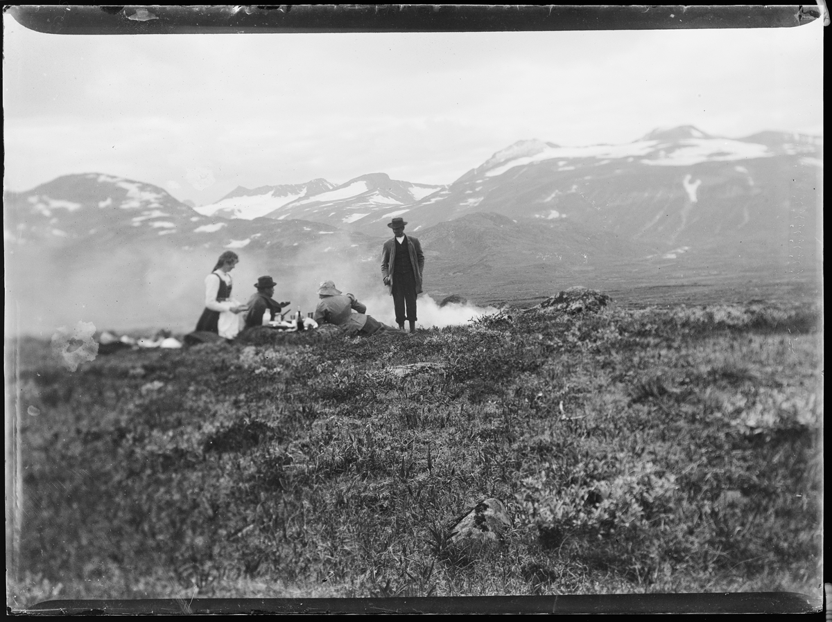 "Heimdalen 1906" (inskr eske). Fra Sjølisæter ved Atnsjøen. Fjellandskap. Snødekte fjell i bakgrunnen. Tre personer ligger i lyngen, en mann står. Det ser ut til at de har tent et bål. Alle er vendt fra kameraet.