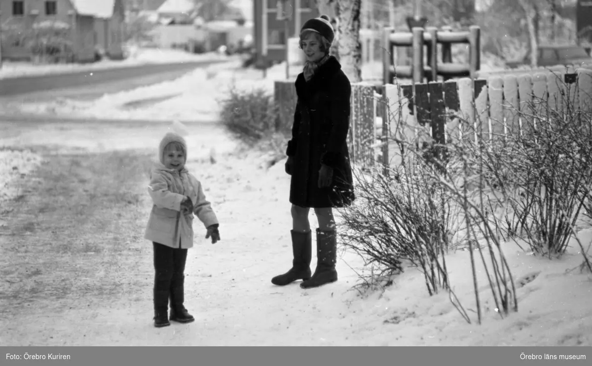 En kvinnas dagbok, Hemmafru Göhle 7 december 1970 

Det står en kvinna i kappa och mössa och en flicka i ljus jacka och mössa på en trottoar.