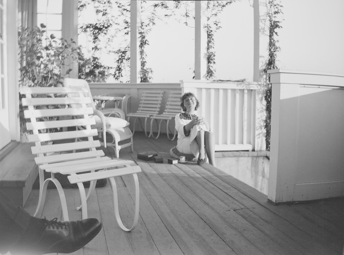 En kvinne sitter øverst på en verandatrapp med kaffe og sigarett. Ved siden av henne på gulvet står noen bokser. Verandaen er møblert med stoler og bord. Klatreplanter henger fra taket. En herrefot med sko sees i det ene hjørnet av bildet. Det er sommer på Linderud Gård.