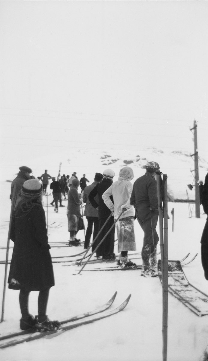 Menn og kvinner står på hver sin side av en bakke eller løype, de fleste med ski på bena. I bakgrunnen sees fjell.
