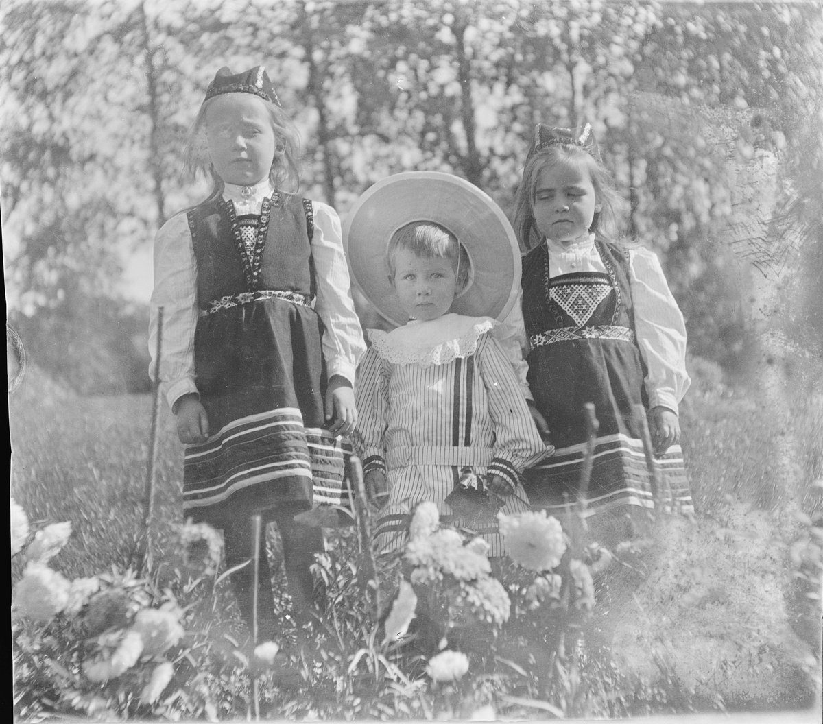 To jenter i hardangerbunad med perlet bringeduk og belte og en gutt står ved et blomsterbed fullt av georginer/dalia.