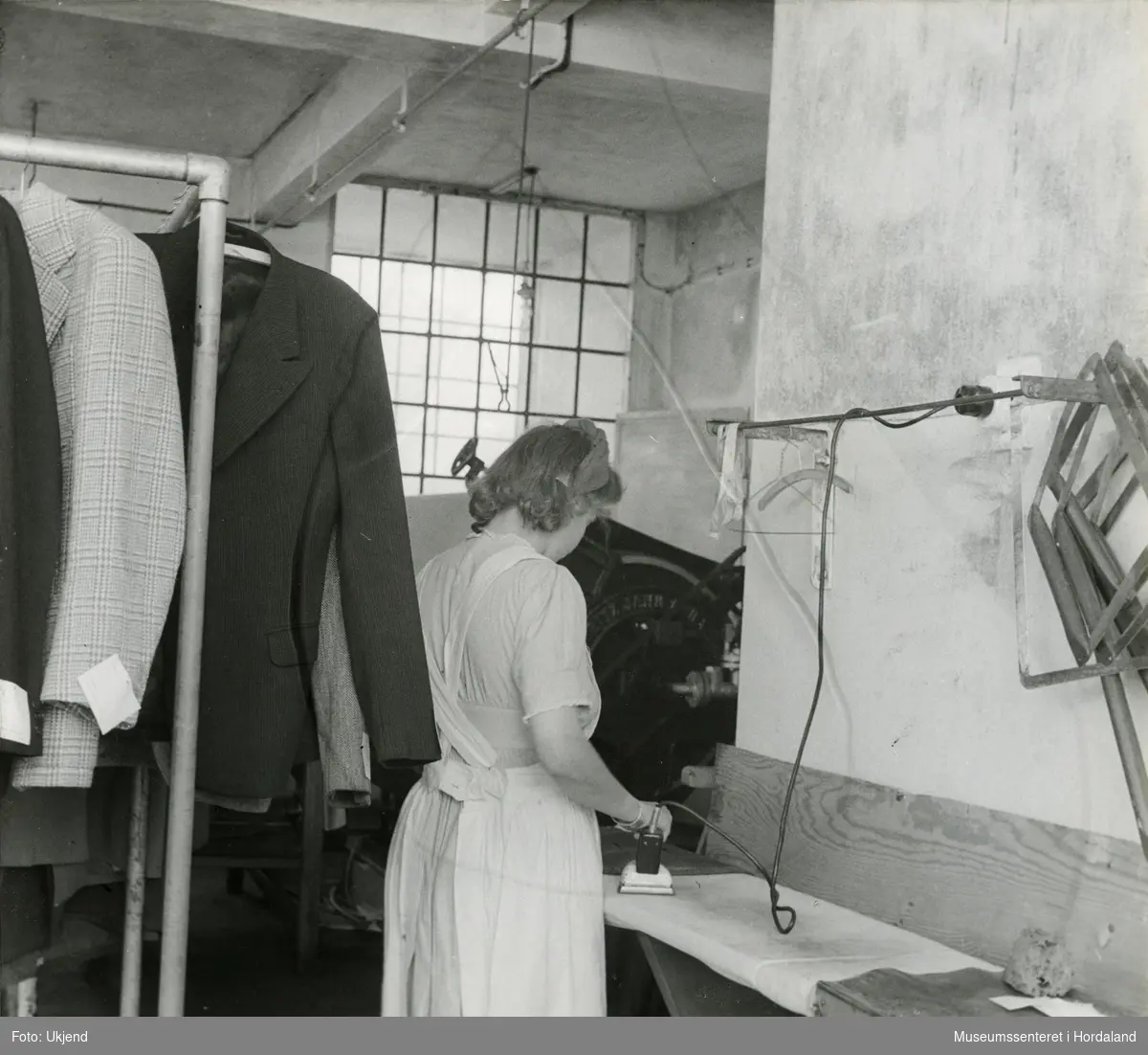 Kvinne stryk klede i presseriet ved Wilhelm Kahrs Kemisk Renseri & Farveri, Bergen. Kalander (valseverk som jamnar ut tekstilen) i bakgrunnen.