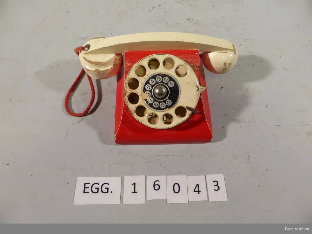 En rød leketelefon med hvitt rør og tallskive. Tallskiven går rundt, røret har vært festet til telefonen med en rød tråd. Tallskiven har nummer fra 0-9.