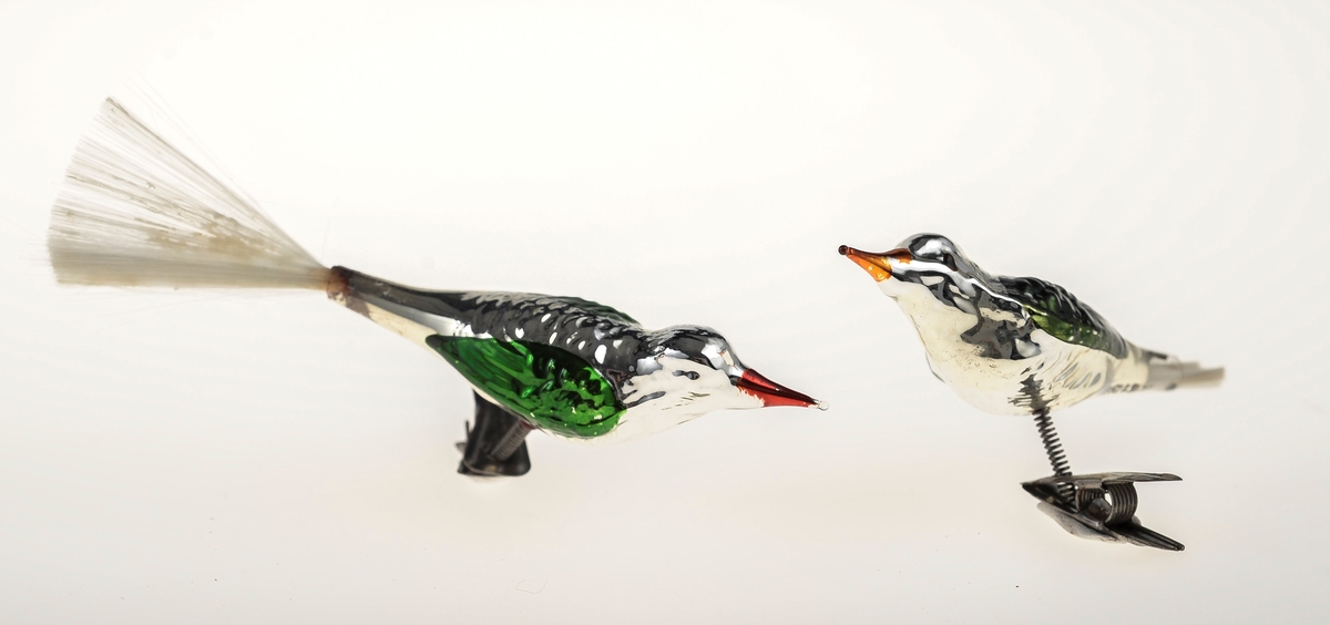 To glassfugler av lik urforming og lik dekor. Fuglene er sølvfarget med rødoransje nebb og grønne vinger. Den ene har stjert av nylonhår, mens det mangler på den andre. Begge er montert på metallklyper med fjæring.