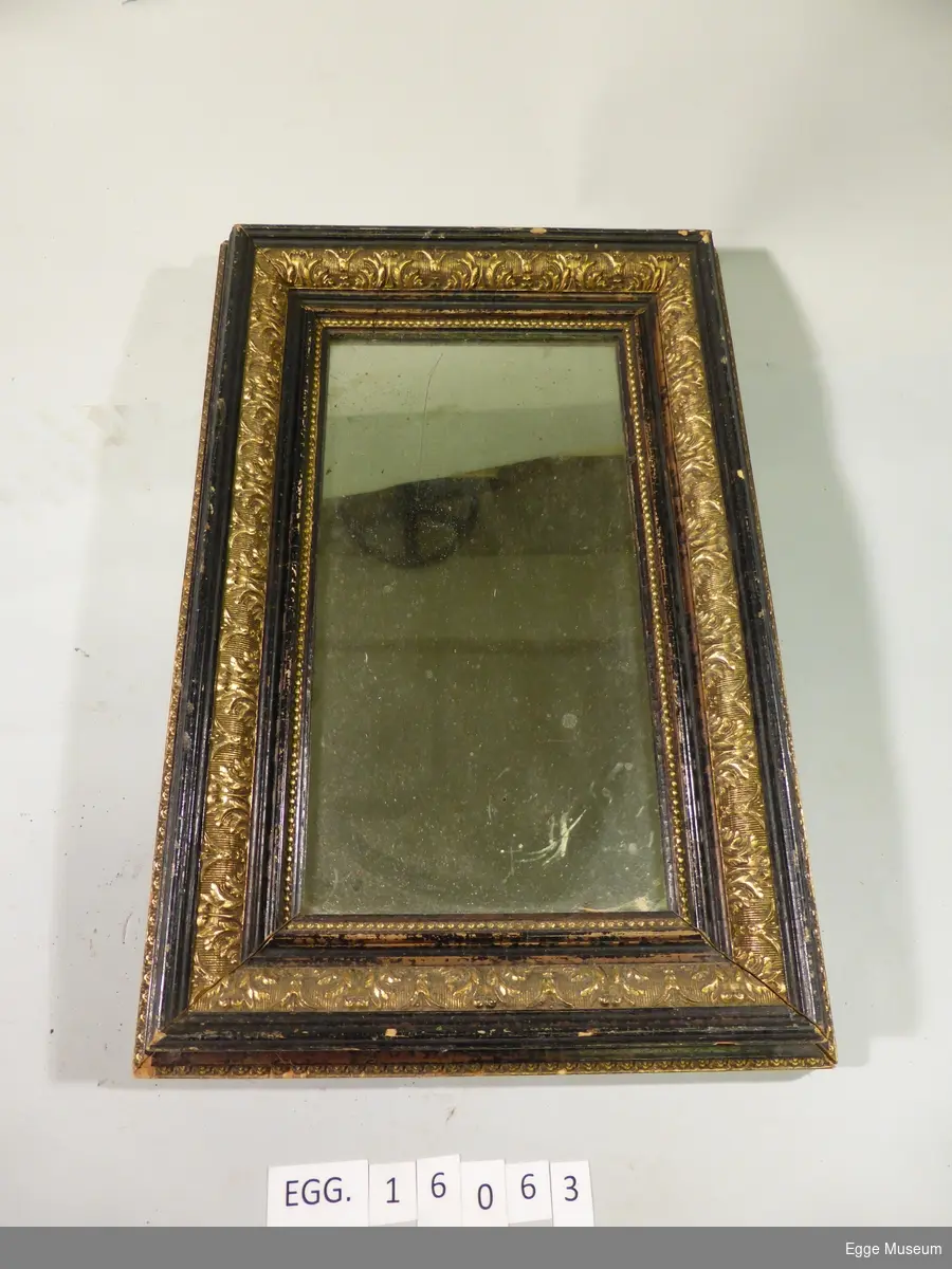 Et rektangulært speil. Rammen er sort med gullforgylte utskjæringer. Innerst mot speilet er det en gullforgylt perlebord. Opphenget er en skru med et øye som er skrudd inn i toppen.

Speielt har et ærverdig preg.
