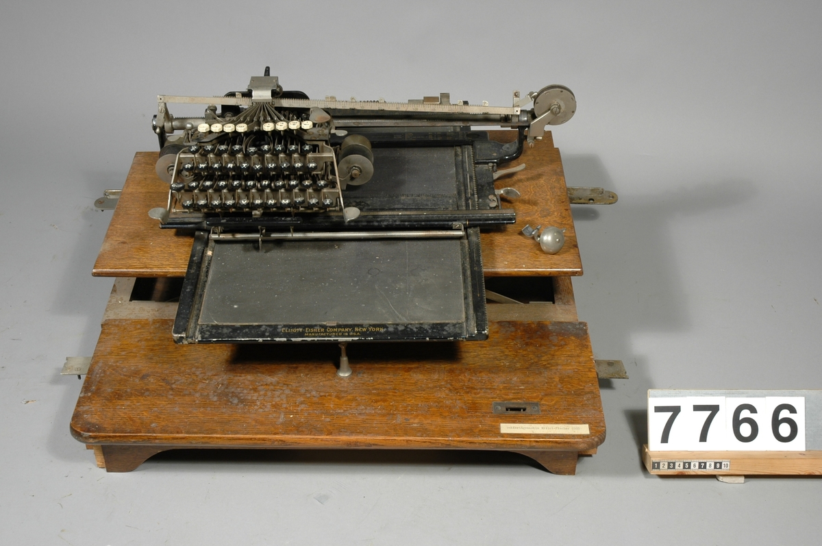 Bokföringsmaskin för skrift i bundna böcker, monterad i mindre jalusiskrivbord. "Elliot-Fisher Company, New York Manufactured in USA", 2 skift, osynlig skrift, kolumnfinnare för enheter t.o.m. milliontal.  På en fastklistrad maskinskriven lapp: "Elliot-Fisher 1905".