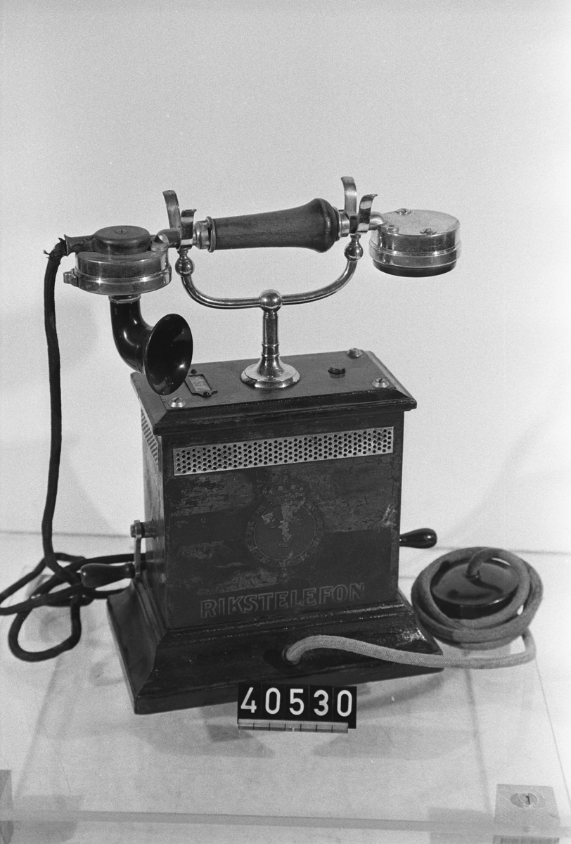 Telefonapparat för lokalbatterisystem tillverkad vid Telgrafverkets Verkstad 1895. Bordsmodell med träbotten, plåtkåpa, textilklädda snören till mikrotelefonen och den extra hörtelefonen samt klyka av metall. Den åtefjädrande tryckomkastaren på kåpans ovansida stänger tillfälligt av mikrofonen vid lyssning under ett samtal för att förbättra hörbarheten i hörtelefonen.