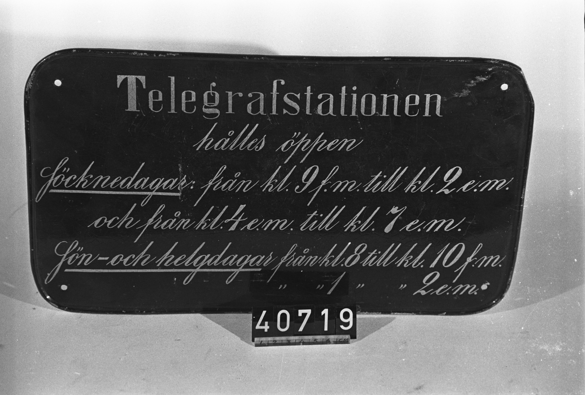 Stationsskylt i svartlackerad plåt, typ "Nr 3". Text: "Telegrafstationen hålles öppen Söcknedagar: från kl. 9 f.m. till kl. 2 e.m. och från kl. 4 e.m. till kl. 7 e.m. Sön- och helgdagar från kl. 8 till kl. 10 f.m. från kl. 1 till kl. 2 e.m."