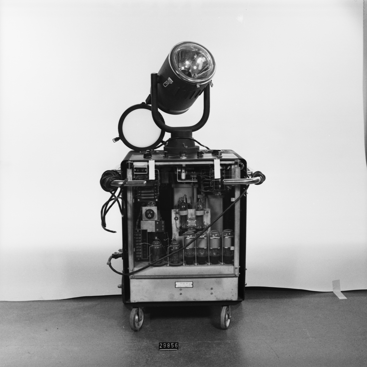 Stroboskop med blixtlampa tillverkad av krymplackerad svartplåt, beslag och handtag i förnicklad metall, monterad på fyra hjul varav två svängbara. Öppen baksida med plexiglas. Märke GM 5500/5501 Nr. 610. V 110-245 W 500. P/S 40-60.