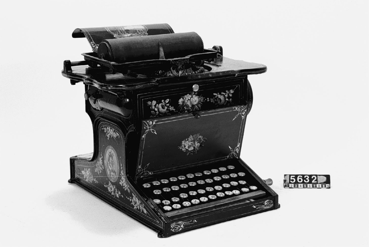 Skrivmaskin märkt: (tillverkad av) "The Sholes & Glidden, Type Writer, Patented, Manufactured by, E. Remington & Sons, Ilion N.Y." (USA). Denna skrivmaskin visar en förbättring av en senare konstruktion omkring 1876 som kom till allmän användning och blev föregångare till de nutida kontorsmaskinerna. Skrivmaskinen är dekorerad med guldblommor och små miniatyrmålningar i färg.