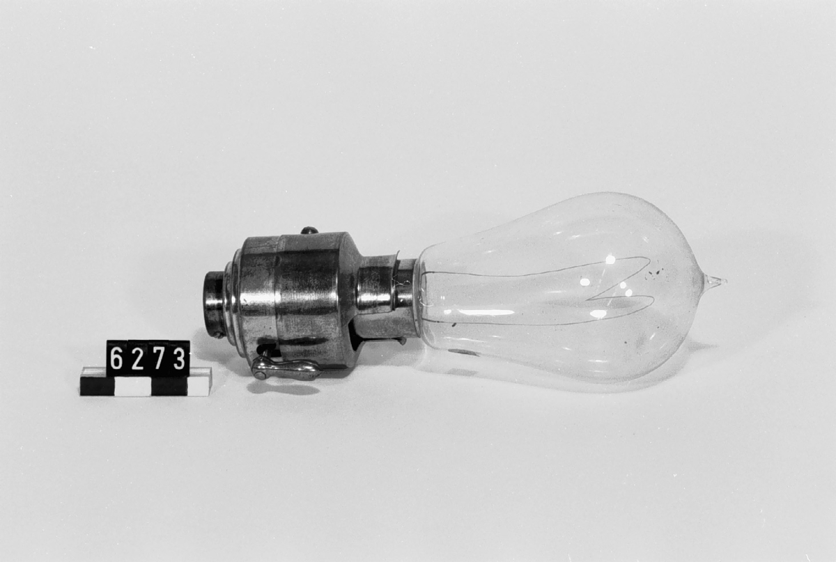 Elektrisk koltrådslampa i hållare med bajonettlås och vridkontakt. Skalet märkt: "Siemens & Halske". Enligt Siemens & Halskes patenterade konstruktion, på 1890-talet mycket spridd i Tyskland. Glödtråd av kolad cellulosa. På lampan etikett: "108".