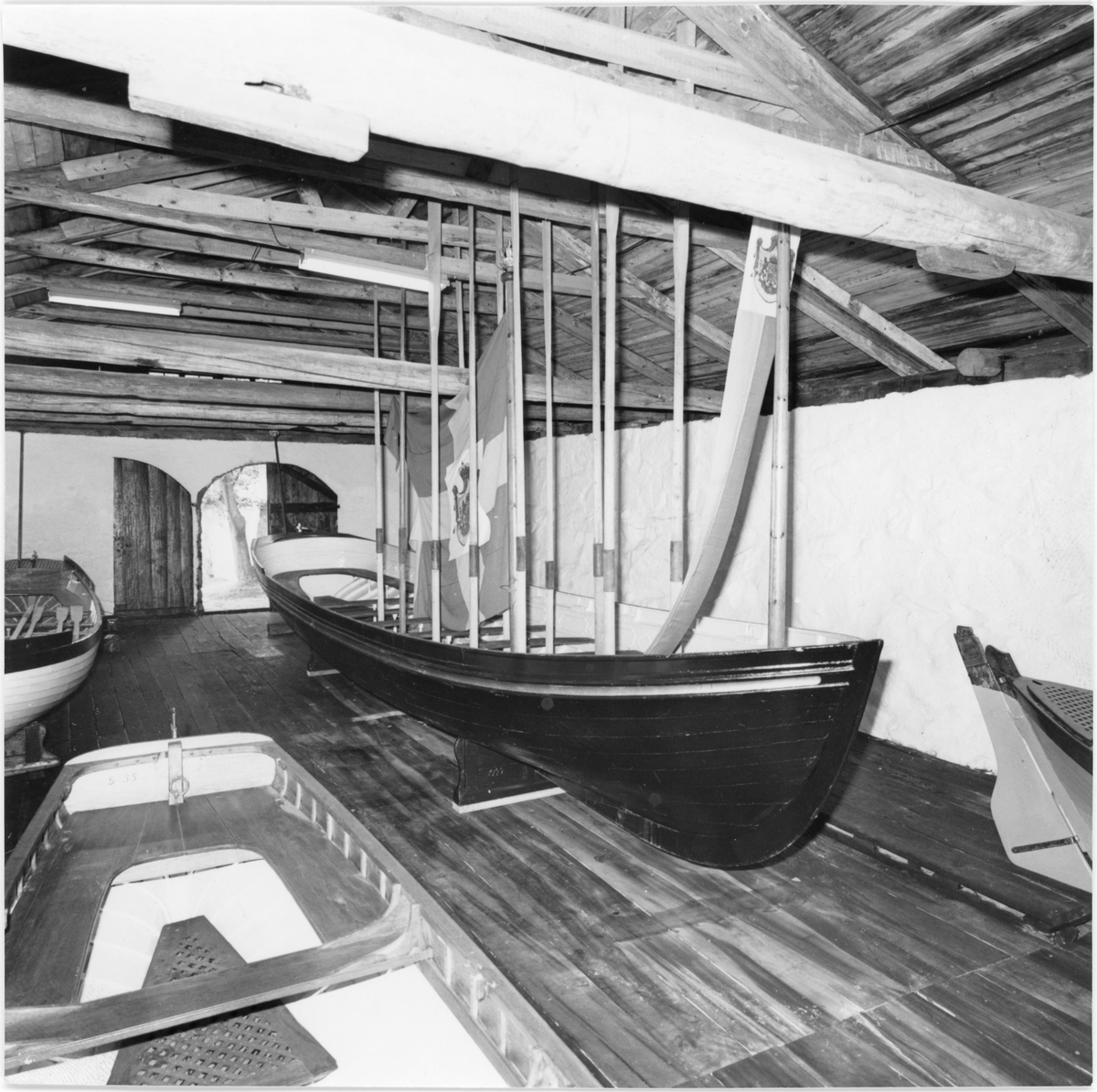 Bilden visar roddbåten kungaslup nr. 44 uppställd i Galjonslundskällaren på Lindholm i Karlskrona. Årorna är uppställd lodrät i båten. Mellan de hänga en flagga. Kungaslupen ingår i en utställning med andra roddbåtar som syns delvis i bilden. Dessutom ser man in i byggnadens takstol.