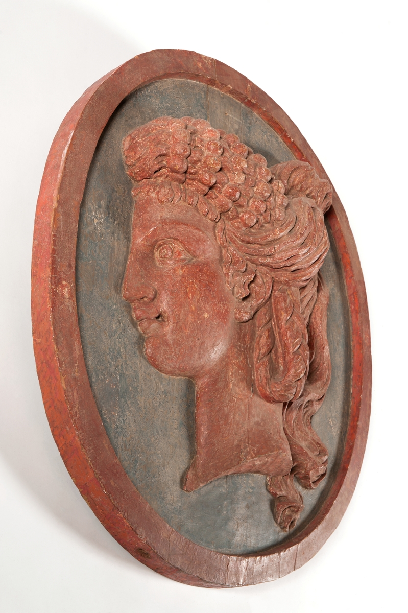 Medaljong, antikiserat kvinnohuvud, enligt uppgift föreställande grevinnan Ehrenswärds huvud. Röd på blågrön botten med oval röd kant.