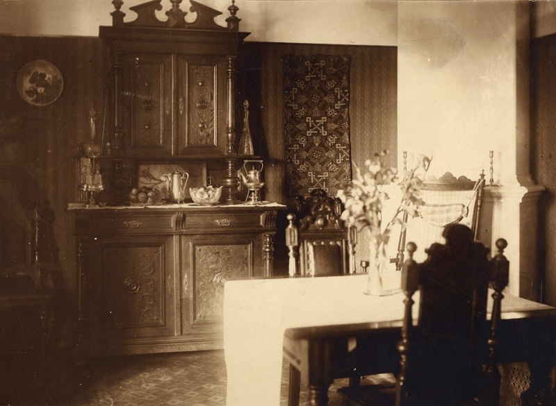 Interiör hög möbel i trä, på väggen hänger ett broderi, i förgrunden till höger matbord och stolar, kakelugn i hörnet, foto: L H Borgström Trelleborg, 77:447.