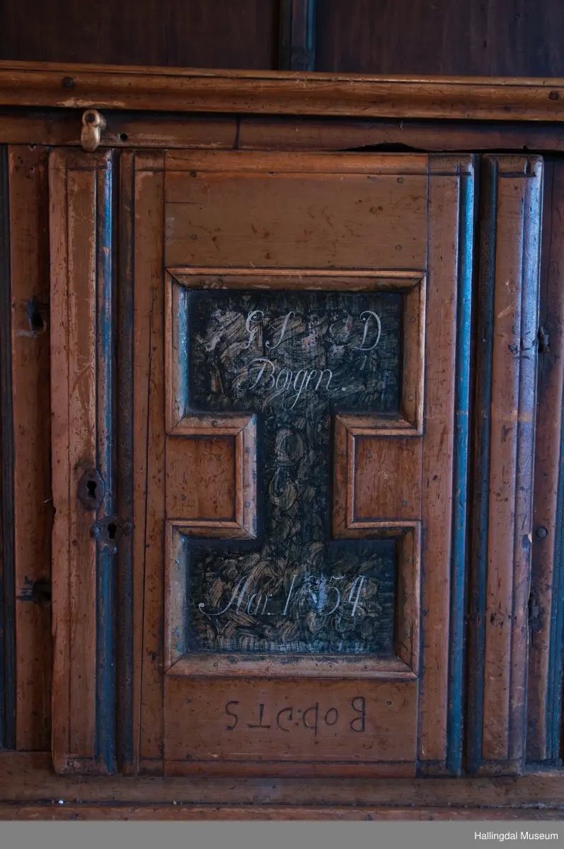 Rester etter rød og blå maling.  Gesims, skråner ned bakover. Profilert. 6 sirkulære hull i arkitraven.  Mellom disse er malt med sorte bokstaver.  Gavlene har utskårne "hylleknekter".  Bakveggen består av fire vertikale planker.  Skjøtene er dekket med profilerte lister.  Bare to biter i nederste del til venstre er fremdeles på plass.  Nederste del er lukket skap.  Tredelt, midterste del er skapdør.  Platen har profillist.  Feltene har speilfyllinger.  Blåmalte.  Klosset marmorering.  Speilfyllingenes form er som en liggen H.  Har profillist.  Skapet er satt på en ny grønmalt plate.  På døren er malt hvite bokstaver.  Nederst på døren er skåret inn bokstaver, opp-ned.  To nøkkelhull m. jernbeslag på venstre side av døren.  Over døren til venstre, en dreibar trekloss, holder igjen.  Innvendig har døren merke etter stort jernbeslag på samme side som nøkkelhullene.  To lister skåret i treverket minner om lange jernbeslag,-smalner mot nøkkelhullene.  Akantusranker i rødt, blått og hvitt, meget avslitt.  En hylle i skapet. Mangler bakvegg.  Karmen har på venstre side spor etter to jernhengsler.  Øverst på høyre kortside, mangler gesimsen en bit, og holdes til sideveggen med en jernkrampe.  Jernkrampe også på venstre side.  Skapplaten mangler list på høyre side.  Venstre side, fra gesimsen og ned har spor etter å ha stått inntil en vegg eller et annet møbel.