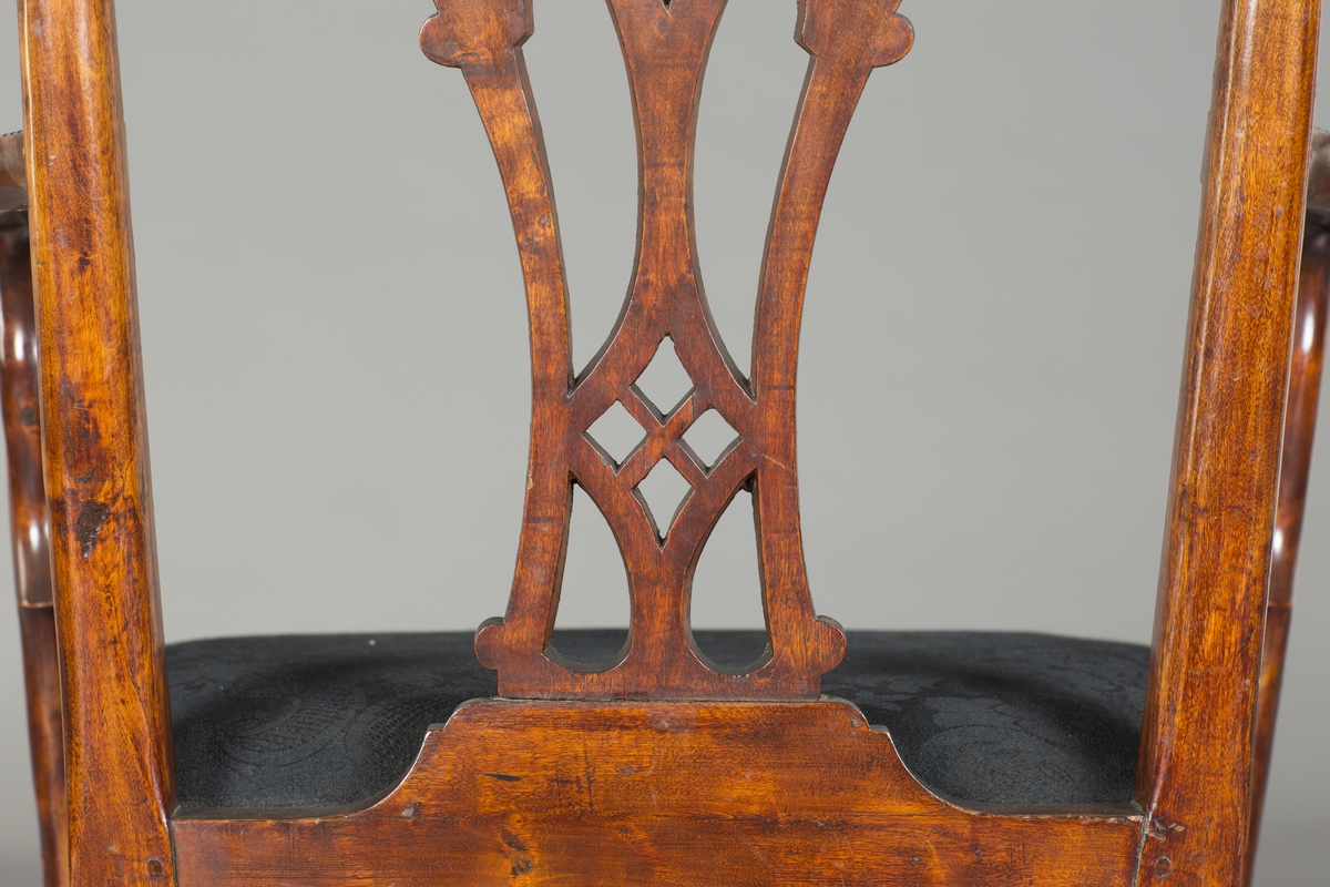 Trestol med svart stoltrekk som ligner på en Chippendale-stol. Stolen har fire firkantede avsmalende ben som er kannelerte. H-formet sprosse mellom bena. Påspikret dekorasjon i hjørnene mellom ben og sarg. Innrisset romertall på innsiden av sargen. Tannsnitt i sargen, armlener og rundt ryggbrettet. Treverket er beiset og man har lakkert over dette. Dekorativt ryggbrett med svungen form og volutter. Buede armlener som svinger utover ved enden, og buede armlenstøtter. Løst sete trukket i svart tekstil, trolig ikke originalt. To stoler mangler sete. To av stolene har lysere treverk og er trolig lagd i en annen tresort. Trolig er stolene beiset med en rødbrun farge slik at de skal imitere edeltre.