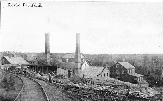 Gammelt bilde fra Klevfos. Togskinner går inn på fabrikkområdet. To store mursteinspiper står blant bygningene.