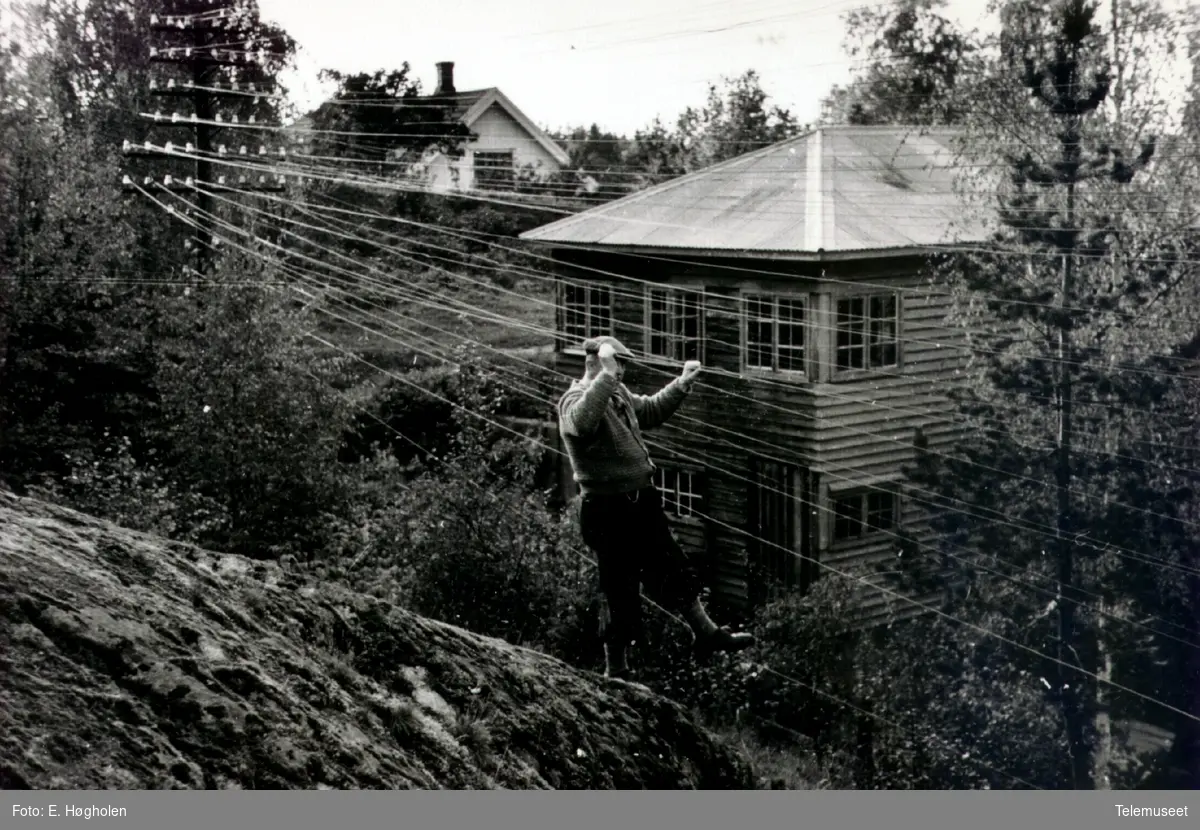 Telefonkurs i skråning med en person oppe i ledningene og et hus (funkis) i bakgrunnen. Dette er på kursen Romedal-Vallset.
