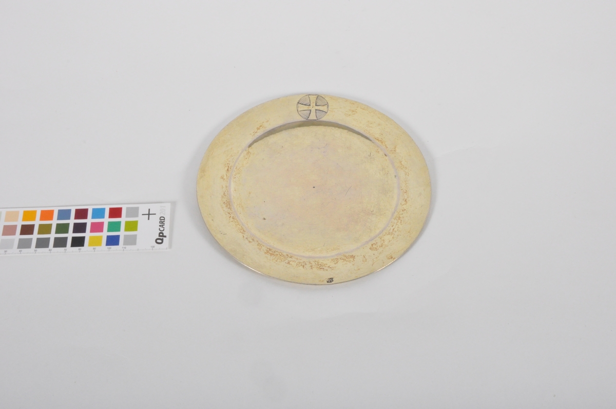Sirkulær disk i sølv. Disken er dekorert med et pattéekors i en sirkel på framsiden, ved kanten.