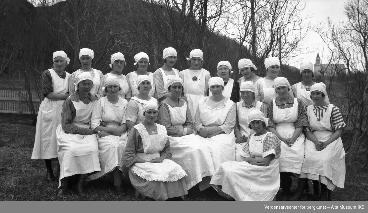 Gruppebilde av 20 kvinner ved husmorskolen i Kåfjord med Kåfjord kirke i bakgrunnen.