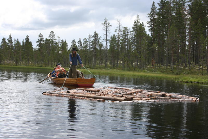 To personer i en robåt som drar en liten tømmerbom etter båten på drevsjøen.