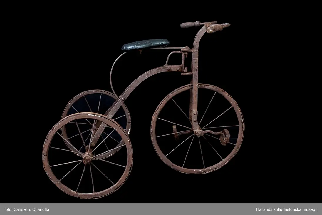 Trehjulig barncykel med metallskodda trähjul. Sadel av galon (i efterhand ditsatt). 