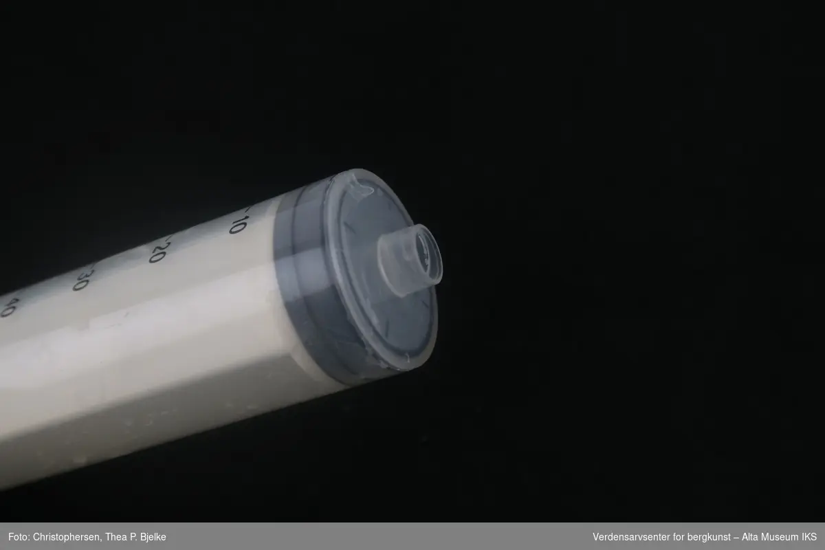 Plastsylinder med grep og trykket gradert skala med målenheter i ml på utsiden, tilhørende plaststempel med svart stempelstopp, og en grå plasthette.  

A: Sprøyten.
B. Ekstradel, hette.