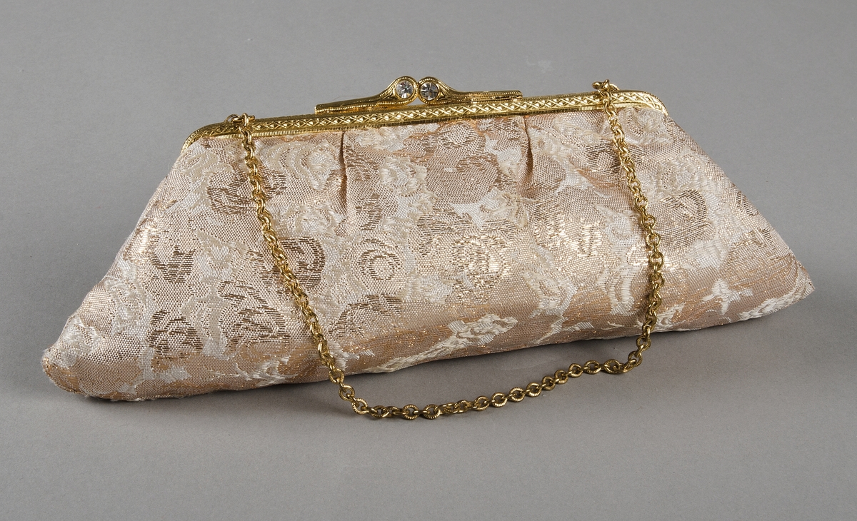 Handväska i guld, vitt och beigebrunt. Handtaget i form av guldfärgad kedja. Två vita stenar i spännet. Ett fack invändigt. 