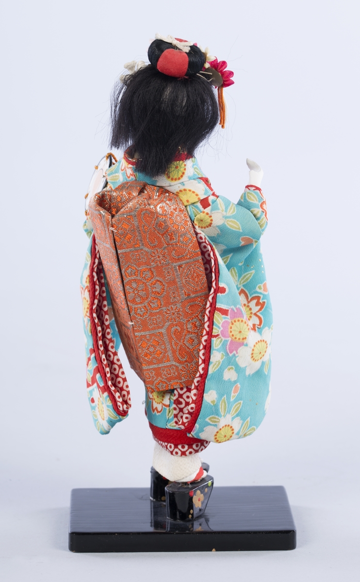Dukke på lav svartlakkert fot. Dukken er ikledd en kimono og geta (høye tresko), og holder en tromme. Blomster i håret.