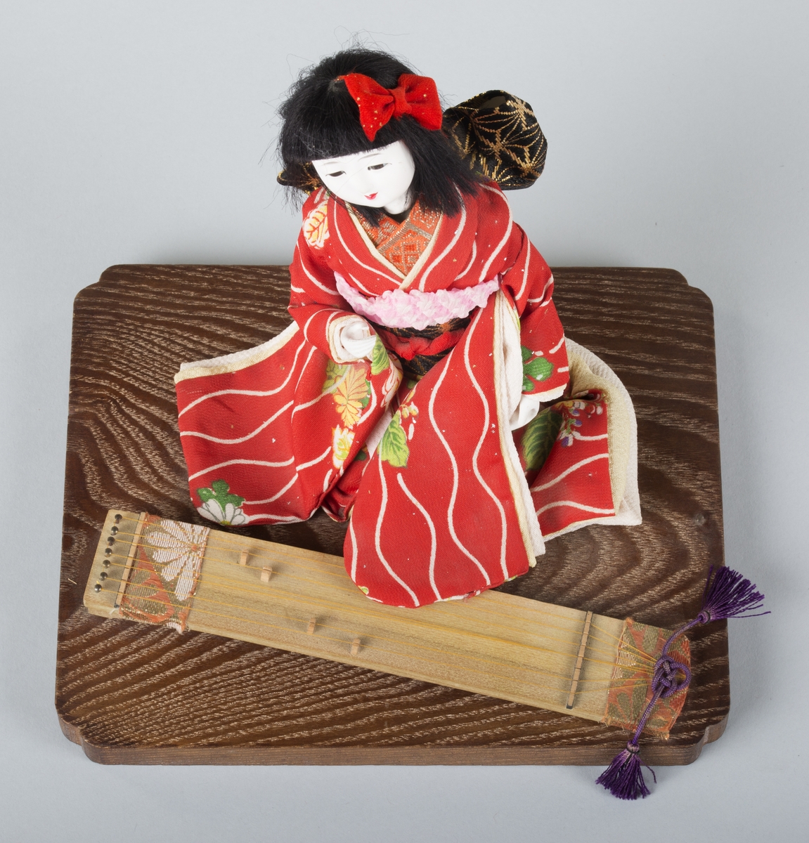 Sittende japansk dukke festet på et lavt platå av tre. Et rektangulært strengeinstrument, koto, foran seg.