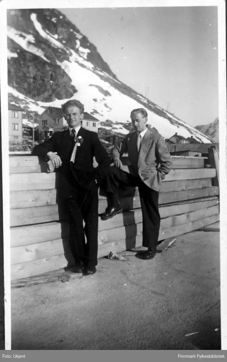 Magnus og Asbjørn Martinsen feirer 17.mai i Øksfjord en gang på 1960-tallet. De to brødrene er kledd i fest klær: dress med slips. De lener seg mot et tregelender og i bakgrunnen kan man se hus og fjell.