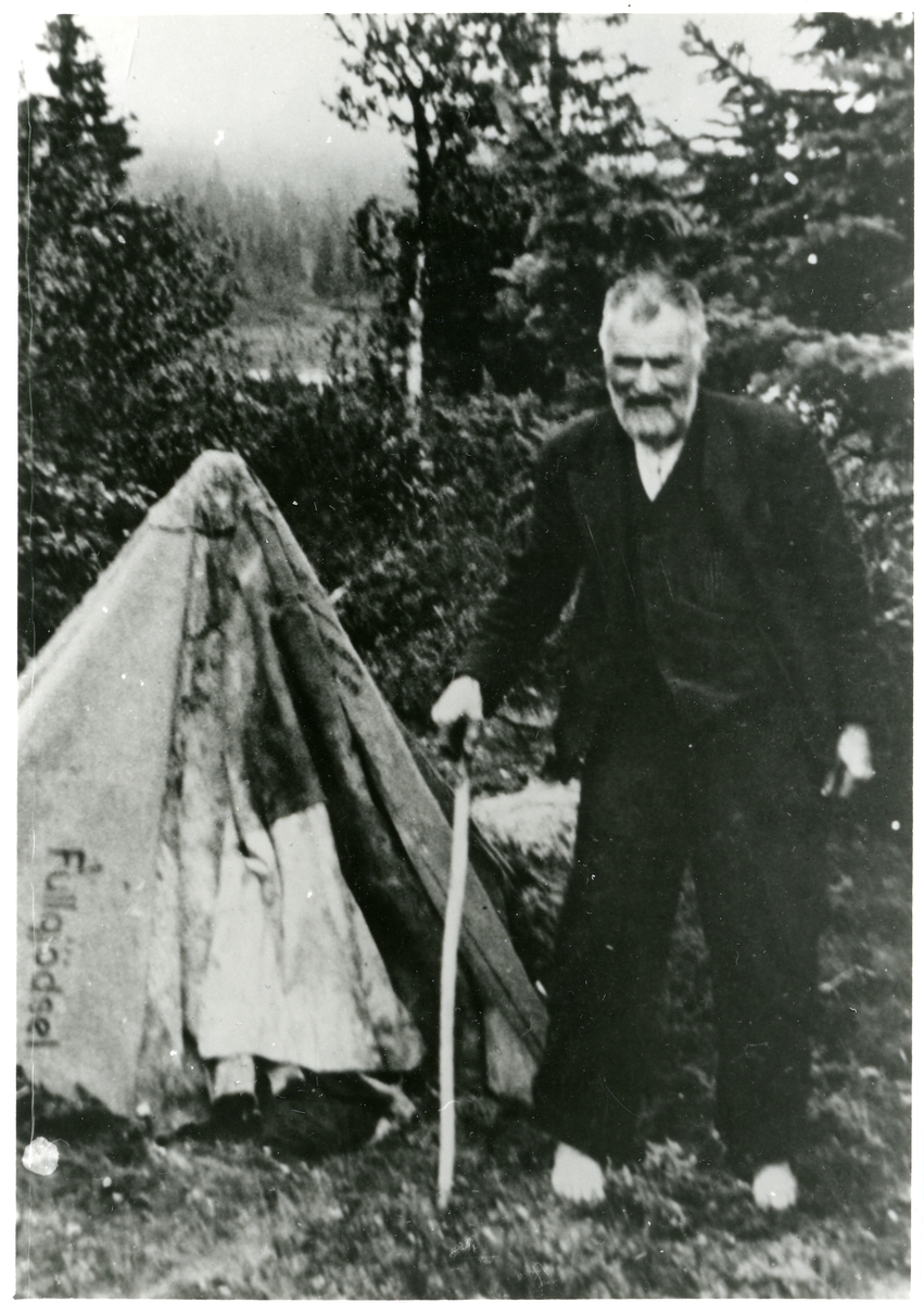 Nub Brager fotografert ute i skogen. Han har en stokk i den ene hånden. I bakgrunnen kan det ses noe som ligner et telt.