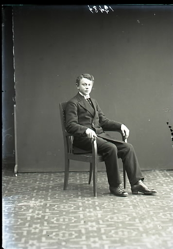 Porträtt ateljéfoto. Bilden föreställer ung man sittande i en stol i helfigur. Beställare: Ivar Nilsén.
