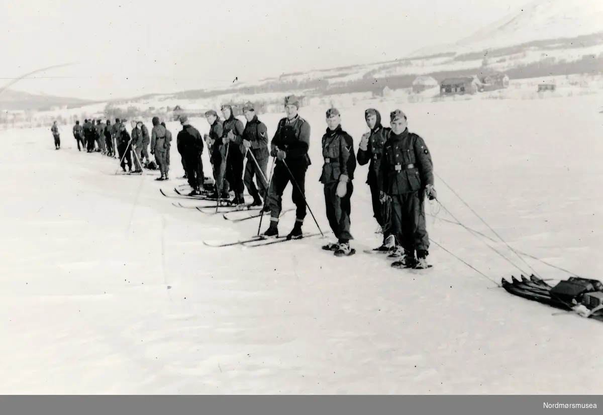 Soldater på ei linje med ski på beina. I bakgrunnen ser vi noen bygninger.