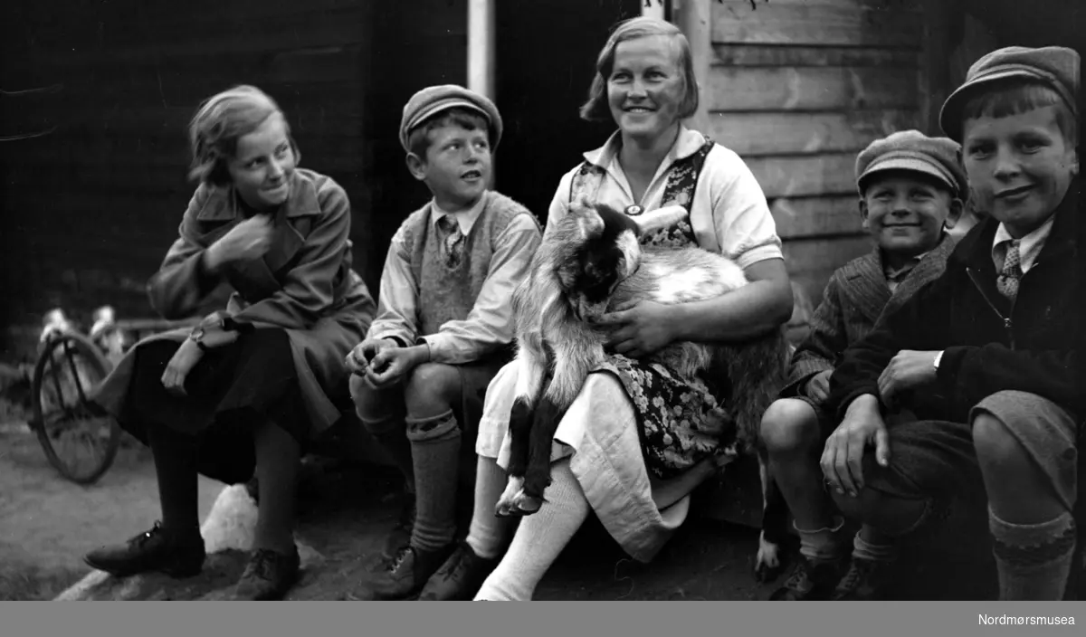 Gruppe av fire barn og en ung kvinne med en geit, sittende utenfor et hus, trolig i Kristiansund. Barna og kvinnen er trolig fra Sverdrup familien/slekten, eventuelt fra deres bekjentskaper. Datering er ukjent, men trolig omkring 1920 til 1939. Fra Nordmøre museums fotosamlinger. Fra Nordmøre museums fotosamlinger. Reg: EFR
