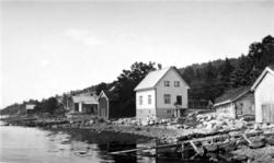 Bygninger ved ei strand. Vi ser bolighus og naust. Huset til