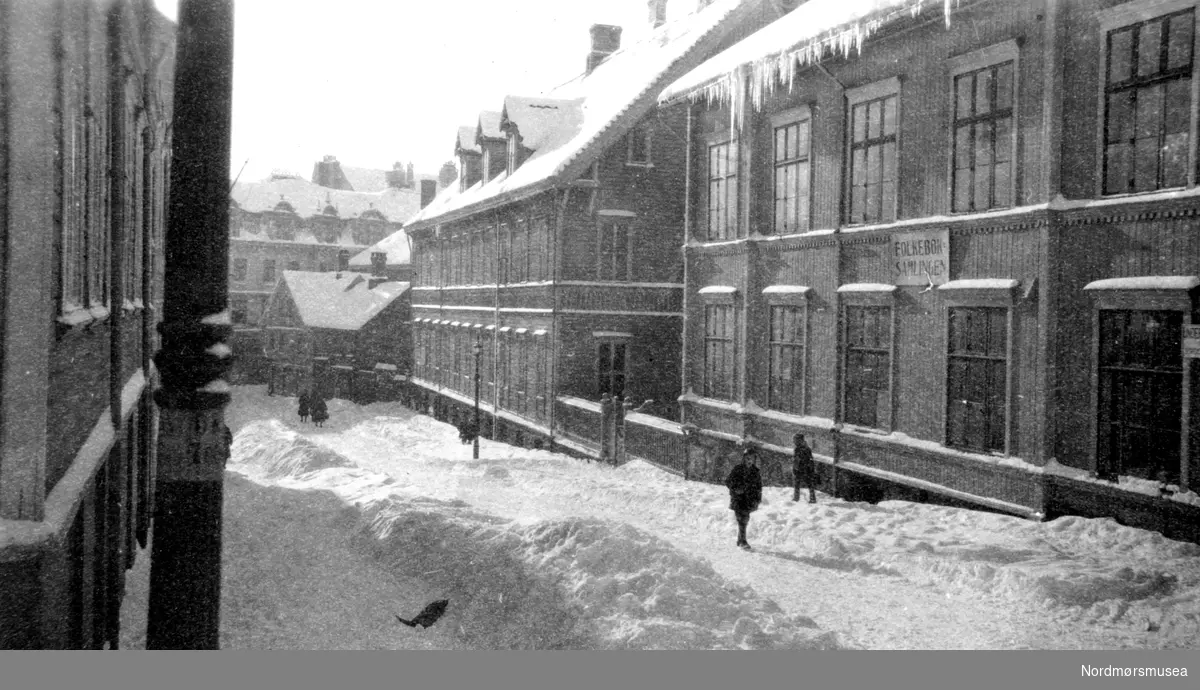 Skilt: Folkeboksamlingen. Biblioteket i Kristiansund, i Nedre Enggate. Enggatens Pikeskole er nabo til venstre. vinter, snø. ca 1880? Fra Nordmøre Museum sin fotosamling.