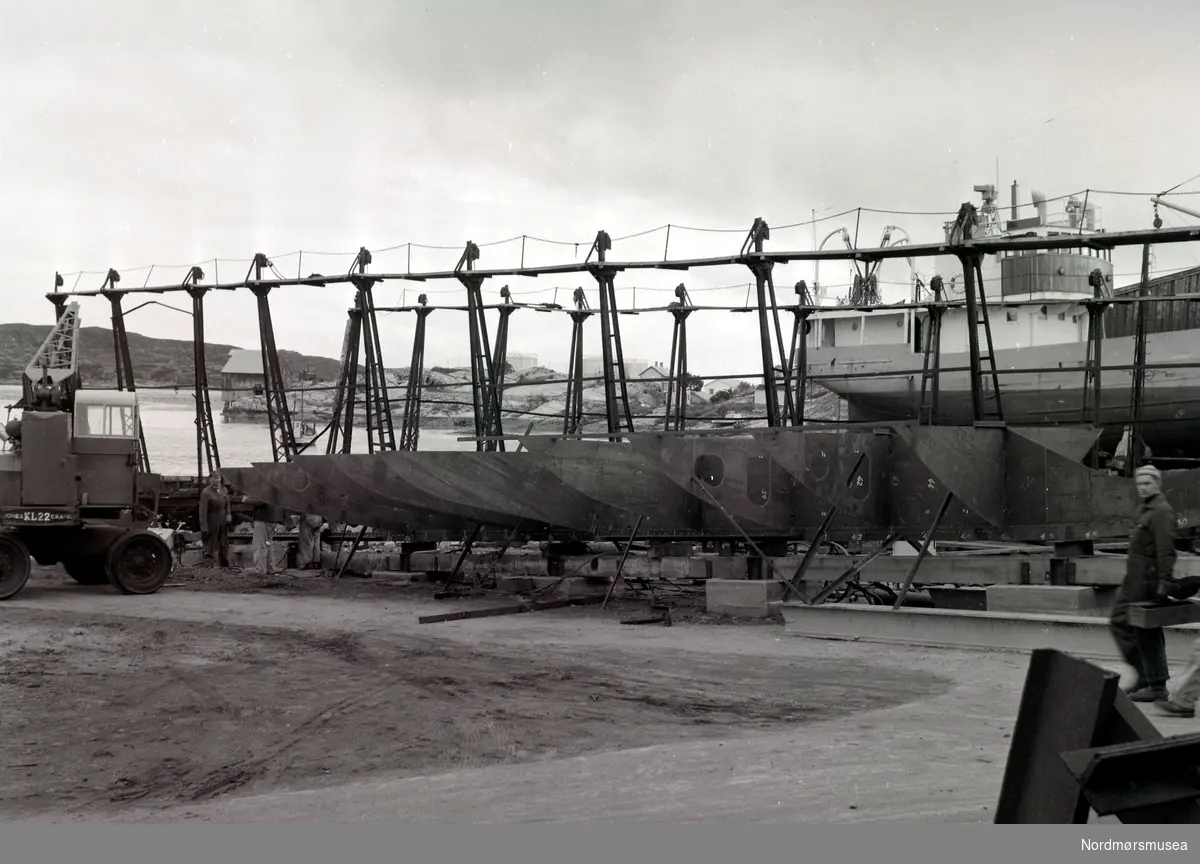 Bildet viser beddingen på Storviks Mek. Verksted på Dahle ved Kristiansund og B/F”Norddalsfjord” verkstedets bnr.14.
På nybygget ser vi montering av bunnstokker.
”Norddalsfjord” ble levert til Møre og Romsdal Fylkesbåtar 15. mars 1961 og hadde følgende hoveddimensjoner: L 31,20 m x B 8,55 m x D 3,35 m og hadde en tonnasje på 159 bruttoregistertonn. Fremdriftsmaskineriet består av 3 Volvo Penta turboladede dieselmotorer type TMD96 på til sammen 420 hk som via kilremdrift var koblet til et felles gir og propellaksel med vribar propell, slik at hver enkelt av motorene kunne kjøres separat. Fergen hadde 2 Bolinders vekselstrømsaggregater type 1052MG på 23 hk hver tilkoblet en generator på 17 kW. Fergen var utstyrt med elektrohydraulisk styremaskin.
Fergen har plass til 18 personbiler og har sertifikat for 160 passasjerer. Forut er det innredet 6 lugarer for offiserer og restaurantpersonale og akterut en mannskapslugar for 4 personer og toppfarten er 11,4 knop og marsjfarten 10,5 knop.
Ferga er verkstedets første nybygg etter B/F”Trygge” som ble levert i 1938.
Ute på sjøen, til høyre for mobilkrana, ses motorskøyta til fisker Olaf Olsen i utlegget.
Personen helt til høyre i bildet er platearbeider Jarl Johannesen og helt til venstre platearbeider Birger Heggemsnes. Helt til høyre ses M/S”Greip” på såpehellingen fra 1901 og foran ”Greip” ses gangveiene på den nye patentslippvogna klinkbygd i stål 1918, den største slippen mellom Bergen og Trondheim.
Til venstre ses verkstedets mobilkran av fabrikat Jones KL22 fra England anskaffet ca. 1959. Den hadde en 1-sylindret dieselmotor og en løfteevne på 2 tonn. I bakgrunnen ses Høttnesset (tidligere kallt Aakvikbergene) og Høttnesbrygga samt tyskerbunkeren og Essos oljetanker og helt til venstre Skorpa. Til høyre for Høttnesbrygga ses røykeriet i tilknytning til brygga. Helt til høyre i bildet ses, nærmest tankene, bolighuset og uthuset til familien Martinus Olsen, senere familiene Olaf og Alfred Olsen, senere familien Olaf Olsen og til høyre for det Essos garasje. Bolighuset og uthuset under hekken på ”Greip” tilhører familien Rikard Kristiansen, senere familien Rikard Rikardsen og motorbåten som står på land foran huset tilhørte sønnen Kåre Rikardsen.
Bildet er fra 1960. Info: Peter Storvik. --
Foto fra Storvik Mekaniske Verksted på Dale, Nordlandet i Kristiansund, hvor vi ser fra arbeidet ved et fartøy på anleggsområdet. . Fra Nordmøre Museums fotosamlinger.