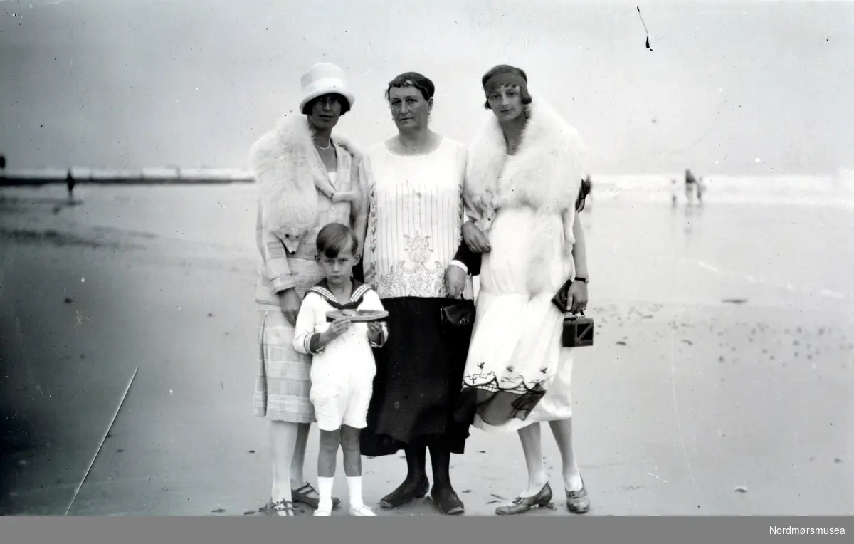 Foto av en gruppe kvinner og en gutt på stranden, ukjent hvor. Kan være i Tyskland eller England, men også andre land er mulige. Datering er usikker, menkan være fra tiden rundt 1920 til 1927. Fra Ida M. Knudtzons fotosamlinger, nå en del av Nordmøre Museums fotosamlinger.
